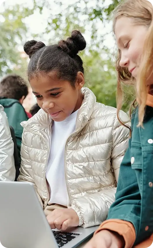Three girls sitting on a bench, using laptops to learn together outside in a sunny environment.