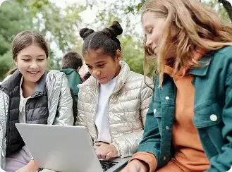 Three girls sitting on a bench, using laptops to learn together outside in a sunny environment.