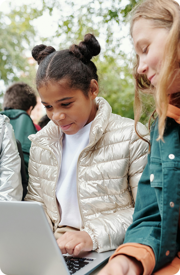 Three girls sitting on a bench, using laptops to learn together outside in a sunny environment.
