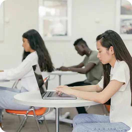 A woman seated at a table, focused on her laptop, with a notebook and pen beside her.