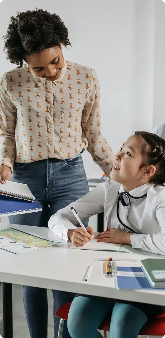 A woman guides a young girl through her homework, sitting together at a table filled with educational materials.