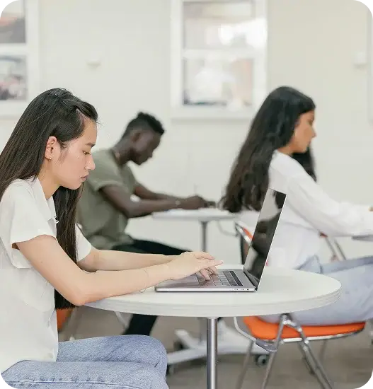 A woman seated at a table, focused on her laptop, with a notebook and pen beside her.