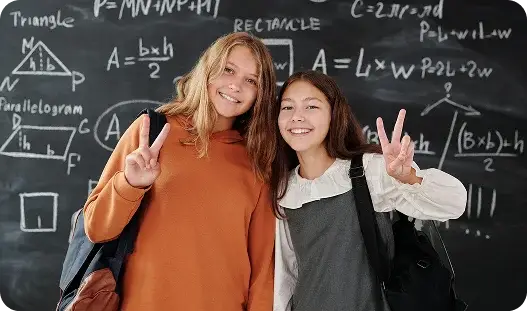 Two girls stand in front of a blackboard filled with various math symbols and equations, engaged in discussion.